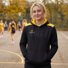Load image into Gallery viewer, Lady model wearing a black and amber Scorpion Sports Heritage Hoodie whilst watching a netball game.