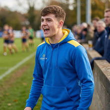 Load image into Gallery viewer, A rugby supporter wearing a Scorpion Sports Heritage Hoodie in royal blue and yellow whilst watching a rugby match from the sideline.
